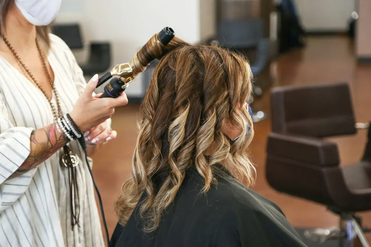 Coiffeuse réalisant des boucles au fer sur une cliente, vue de dos